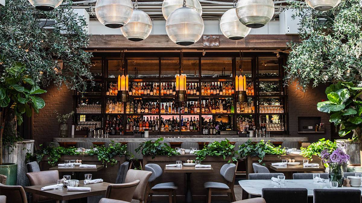 A dining area with grey chairs and wooden tables with lots of greenery surround it and a bar in the background with large light fixtures and a wall of bottle at Aba Restaurant in Chicago, Illinois, USA